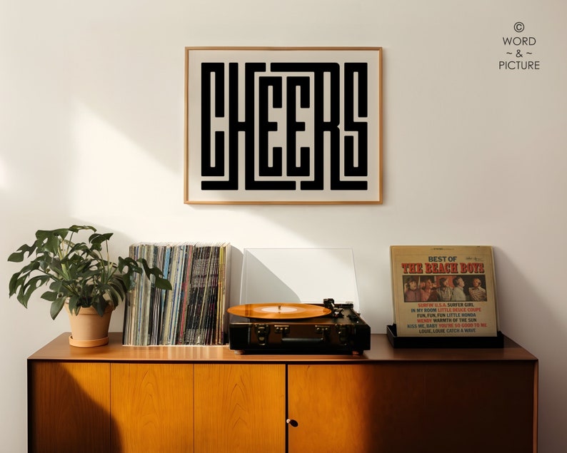 May include: A black and white print with the word "CHEERS" in a bold, geometric font. The print is framed in a thin, black frame and is hanging on a white wall. The print is above a wooden cabinet with a record player and a stack of vinyl records.
