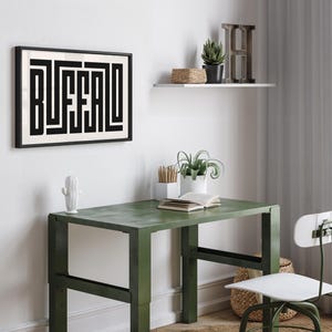 May include: A black framed print with the word "Buffalo" in a geometric design, hanging on a white wall above a green metal desk with a white chair. There are plants and a book on the desk.