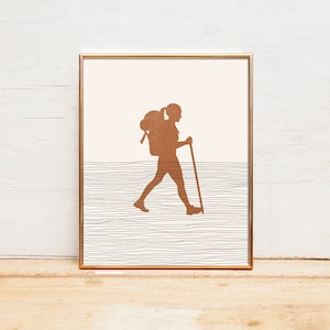 May include: A framed print of a silhouette of a person hiking with a backpack and walking stick. The background is a light pink with a textured gray and white pattern.