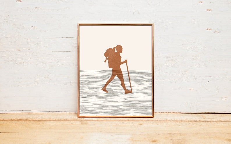 May include: A framed print of a silhouette of a person hiking with a backpack and walking stick. The background is a light pink with a textured gray and white pattern.