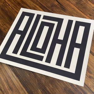 K&ouml;nnte beinhalten: Eine wei&szlig;e Karte mit dem Wort "ALOHA" in schwarzer Schrift, in einem geometrischen Design.