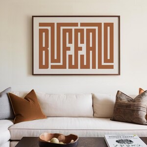 May include: A white couch with brown throw pillows and a brown framed print with the word "BUFFALO" in a geometric design. The print is hanging on a white wall above the couch.