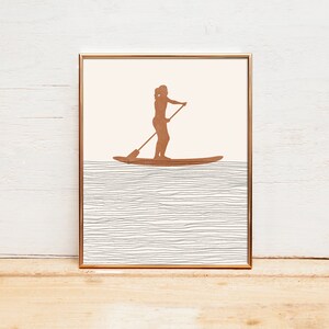 May include: A framed print of a silhouette of a person stand-up paddleboarding on a body of water. The water is represented by a series of thin, gray lines.