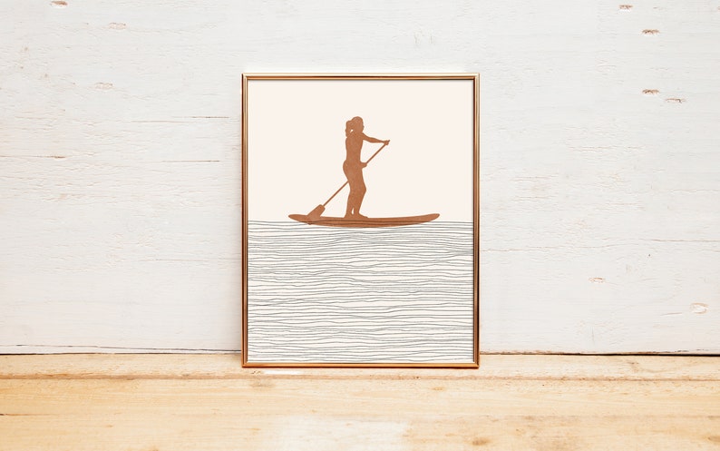 May include: A framed print of a silhouette of a person stand-up paddleboarding on a body of water. The water is represented by a series of thin, gray lines.