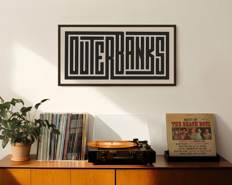 May include: A framed black and white print with the word "OUTERBANKS" in a geometric font. The print is hanging on a white wall above a wooden shelf with a record player, a stack of vinyl records, and a record album by The Beach Boys.