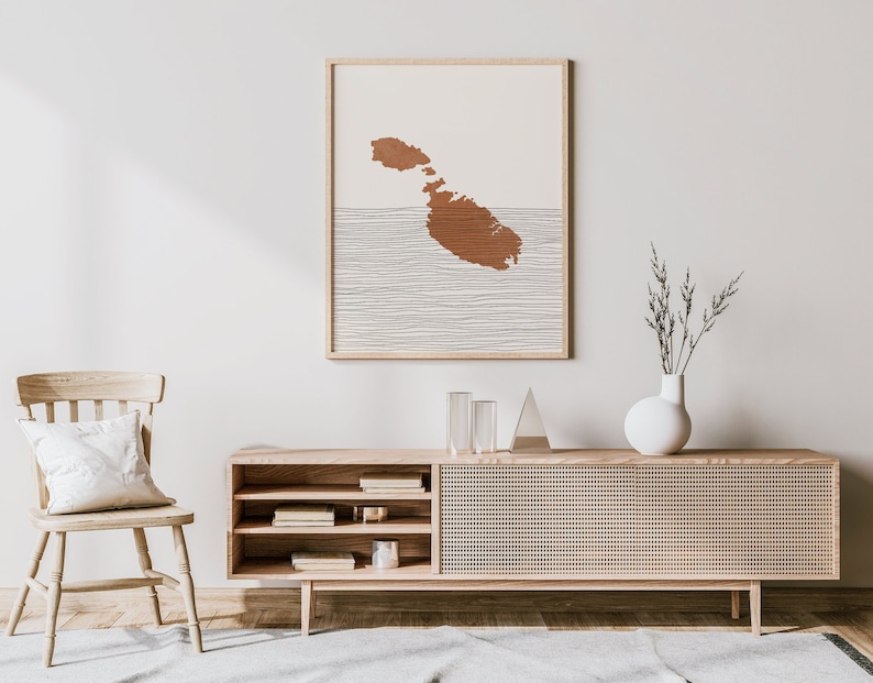 May include: A light wood media console with a woven rattan front, open shelves, and a white vase with dried branches on top. A framed print of the island of Malta in brown and white hangs on the wall behind the console.