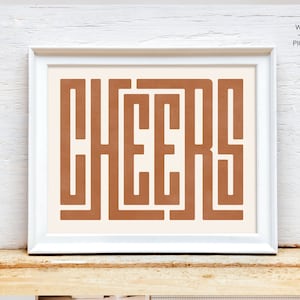 May include: A white framed print with the word "CHEERS" in a brown, geometric font on a cream background.