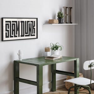 May include: A black framed print with the word "SLAM DUNK" in white letters. The print is hanging on a white wall above a green metal desk with a white chair. There are plants and a wooden shelf with a wooden letter H on the wall.