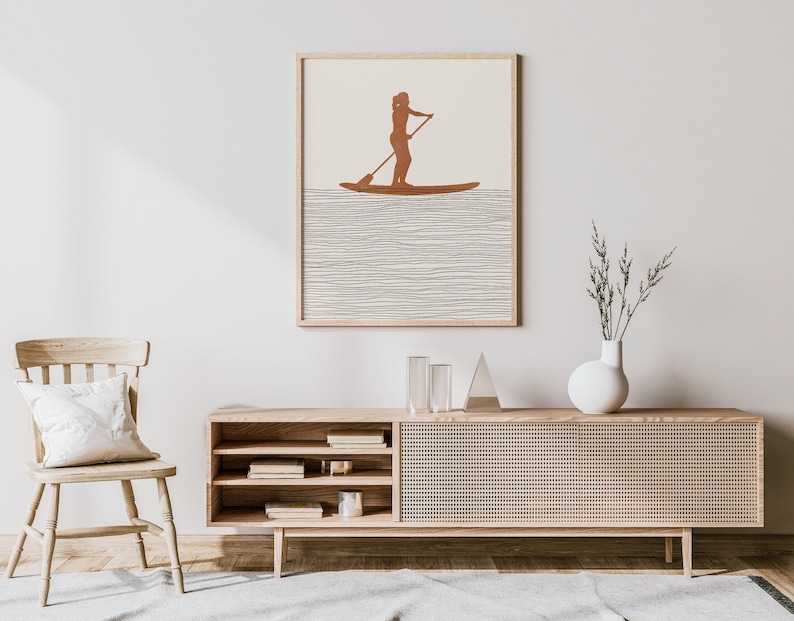 May include: A minimalist illustration of a person paddleboarding on a body of water. The illustration is in shades of brown and white and is framed in a light wood frame. The frame is hanging on a white wall above a wooden cabinet with a woven rattan front.