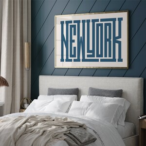 May include: A white bed with a beige blanket and white pillows in a bedroom with a blue wall and a gold framed print that says "NEW YORK" in blue letters.