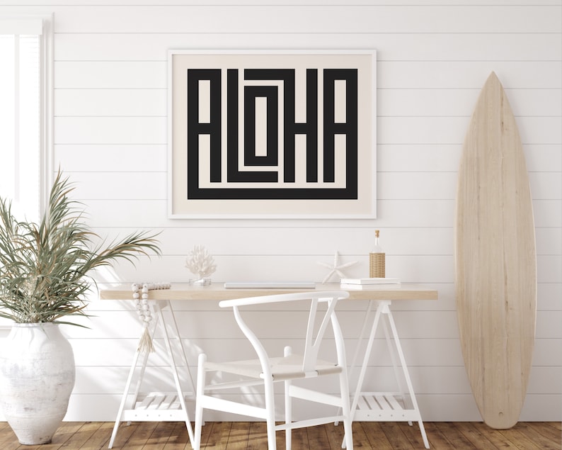 Peut inclure: Une impression encadr&eacute;e en noir et blanc avec le mot "ALOHA" dans un design g&eacute;om&eacute;trique. L'impression est accroch&eacute;e &agrave; un mur blanc en lambris au-dessus d'un bureau blanc avec une chaise blanche. Une planche de surf est appuy&eacute;e contre le mur &agrave; droite de l'impression.