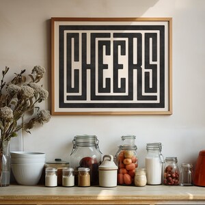 May include: A framed black and white print with the word "CHEERS" in a geometric design. The print is hanging on a white wall above a countertop with various glass jars and bowls.