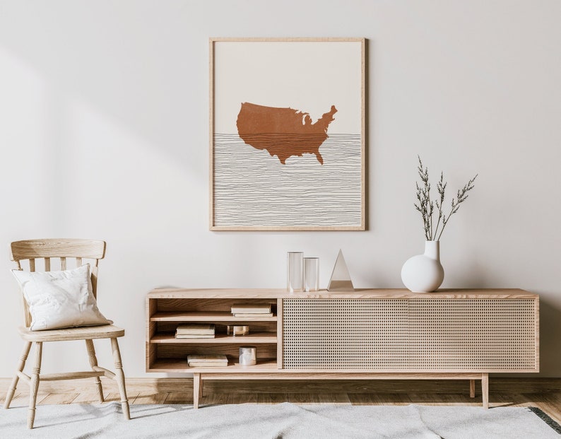 May include: A wooden console table with a woven rattan door and open shelves. A white vase with dried branches sits on top of the table, and a framed print of a brown outline of the United States with white lines is hanging on the wall behind the table.