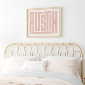 K&ouml;nnte beinhalten: Ein gerahmter Druck mit dem Namen "AUSTIN" in einem rosafarbenen geometrischen Design. Der Druck h&auml;ngt an einer wei&szlig;en Wand &uuml;ber einem hellbraunen Korbkopfteil.