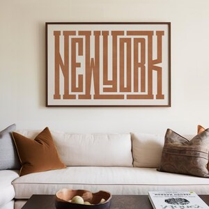 May include: A white couch with brown, gray, and orange throw pillows in front of a white wall with a framed print that says "NEW YORK" in brown letters.