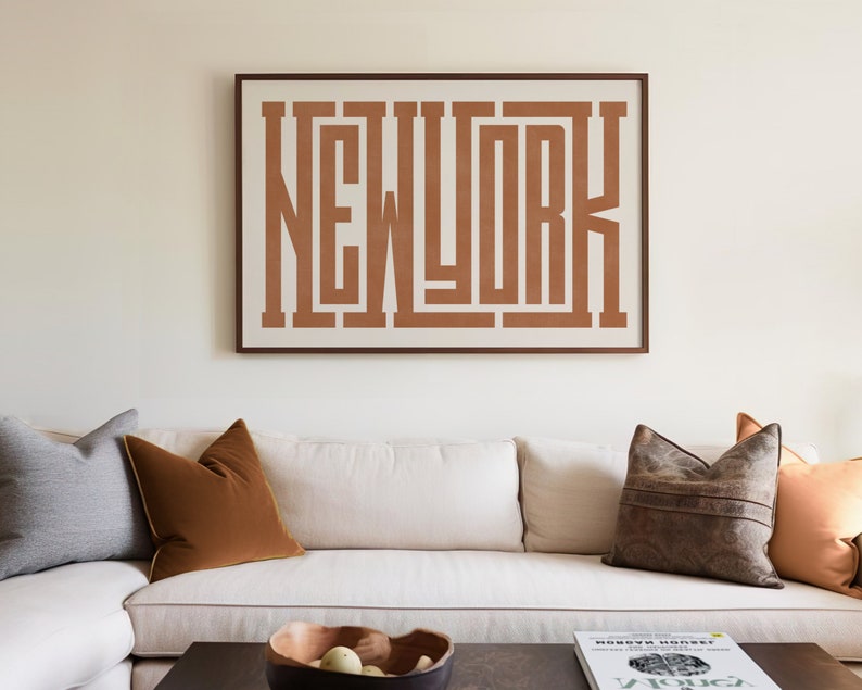 May include: A white couch with brown, gray, and orange throw pillows in front of a white wall with a framed print that says "NEW YORK" in brown letters.
