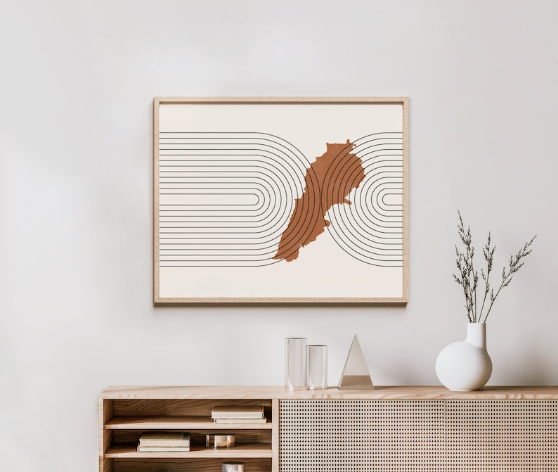 May include: A minimalist art print featuring a brown outline of Lebanon on a white background with black curved lines. The print is framed in a light wood frame and is hanging on a white wall.