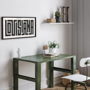 Puede incluir: Una impresi&oacute;n enmarcada en blanco y negro con la palabra "LOUISIANA" en un dise&ntilde;o geom&eacute;trico. La impresi&oacute;n est&aacute; colgada en una pared blanca sobre un escritorio de metal verde con una silla blanca. Hay plantas y un estante con una letra H de madera en la pared.