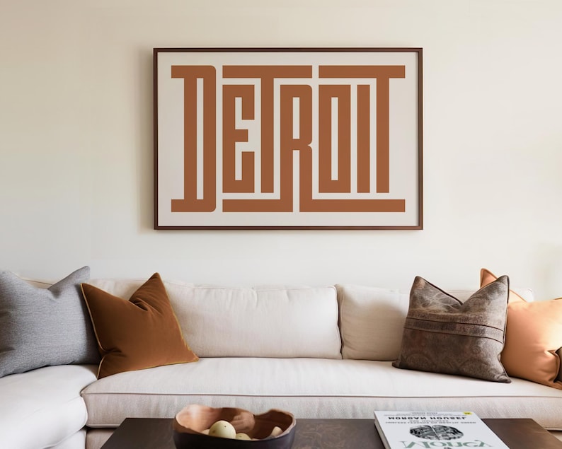 May include: A white couch with brown throw pillows in a living room. A framed print with the word "DETROIT" in brown letters is hanging on the wall above the couch.