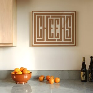 May include: A kitchen counter with a brown bowl filled with oranges, two bottles of wine, and a framed print with the word "CHEERS" in a brown geometric design.