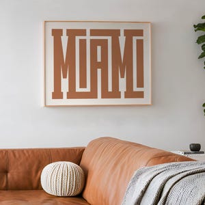 K&ouml;nnte beinhalten: Ein gerahmter Druck mit dem Wort "MIAMI" in brauner Farbe, wobei sich die Buchstaben &uuml;berlappen. Der Druck h&auml;ngt an einer wei&szlig;en Wand &uuml;ber einem braunen Ledersofa.