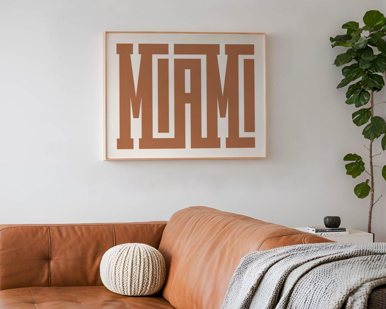 K&ouml;nnte beinhalten: Ein gerahmter Druck mit dem Wort "MIAMI" in brauner Farbe, wobei sich die Buchstaben &uuml;berlappen. Der Druck h&auml;ngt an einer wei&szlig;en Wand &uuml;ber einem braunen Ledersofa.
