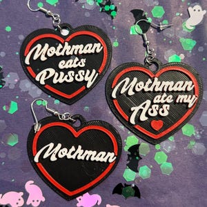 May include: Three heart-shaped earrings with black bases and red borders. One earring features the white text "Mothman eats Pussy", another says "Mothman ate my Ass" with a small red heart, and the third displays the white text "Mothman".