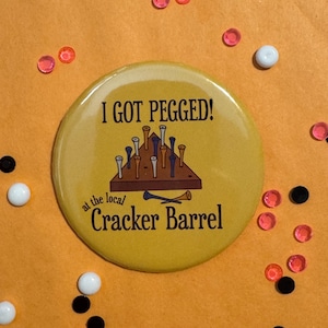 May include: A round yellow button featuring a graphic of a triangular peg game board with various colored pegs. The text reads I GOT PEGGED! at the local Cracker Barrel in black lettering. The button is surrounded by small scattered beads.