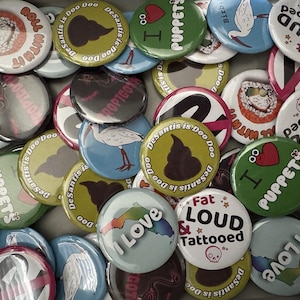 May include: A collection of round pin-back buttons featuring various designs including a brown poop icon with the text DeSantis is Doo Doo, a red heart with I Love Puppets, a white button with Fat LOUD & Tattooed, and sushi illustrations.