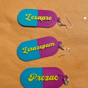 May include: Three pill-shaped earrings featuring a split design of bright blue and purple. Each earring has yellow raised lettering spelling out Lexapro, Lorazepam, and Prozac respectively. The earrings include silver-toned metal hooks for attachment.
