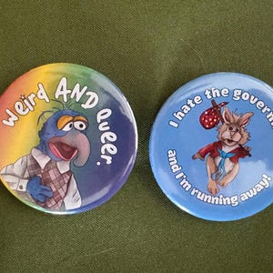 May include: Two circular pinback buttons. The left button features a blue character with a large nose, wearing a plaid vest, with the text "Weird AND Queer." The right button features a rabbit carrying a red polka-dot bindle, with the text "I hate the government and I'm running away!"