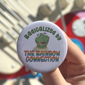 May include: A circular pin-back button with a white background featuring a green frog illustration in the center. The text reads Radicalized by in green letters above the frog and THE RAINBOW CONNECTION in rainbow-colored letters below.