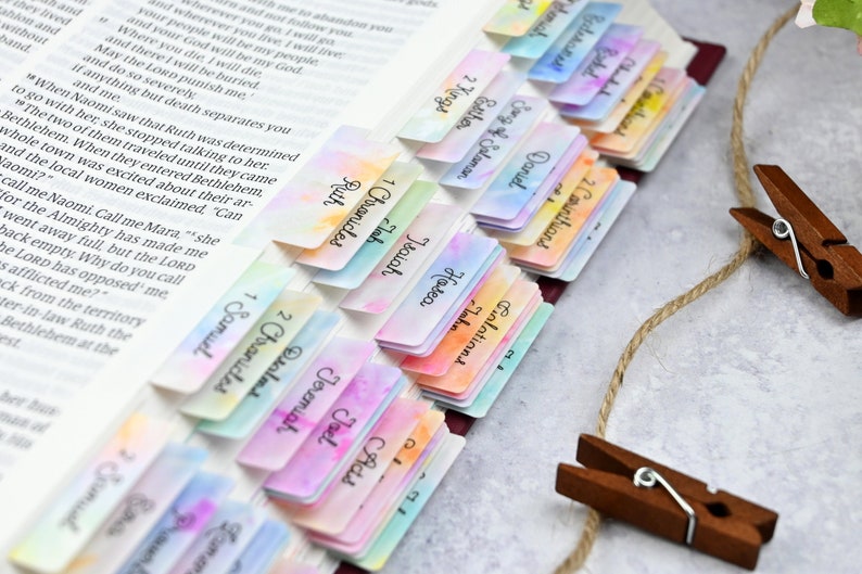 May include: A close-up of a Bible with colourful, watercolour-painted bookmarks. The bookmarks are labelled with different books of the Bible, including Ruth, 1 Samuel, 1 Chronicles, 2 Chronicles, Psalms, Isaiah, Jeremiah, Ezekiel, Daniel, Hosea, Joel, Amos, Obadiah, Jonah, Micah, Nahum, Habakkuk, Zephaniah, Haggai, Zechariah, Malachi, and Song of Solomon.