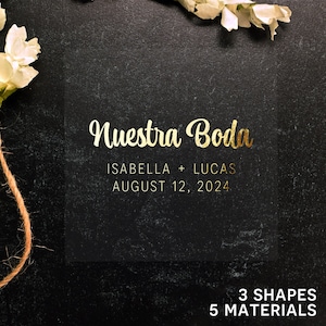 May include: A black chalkboard with gold lettering announcing a wedding. The text reads "Nuestra Boda" with the names Isabella and Lucas and the date August 12, 2024. The chalkboard is surrounded by white flowers and twine.
