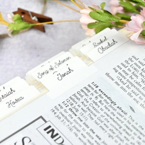 May include: Close-up of a white, leather-bound book with tabs for different sections of the Bible. The tabs are labeled with the names of books of the Bible, including Isaiah, Hosea, Song of Solomon, Jonah, Ezekiel, and Obadiah. The book is open to the book of Jonah, with the text "Jonah 1:10" visible.