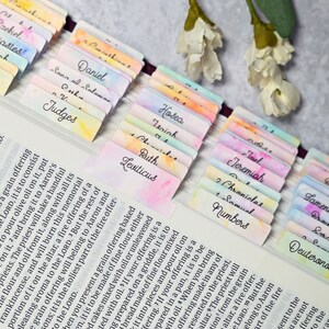 May include: A set of colourful bookmarks with the names of books of the Bible written in black ink. The bookmarks are arranged in a row, with the names of the books visible. The bookmarks are made of paper and have a watercolour effect.
