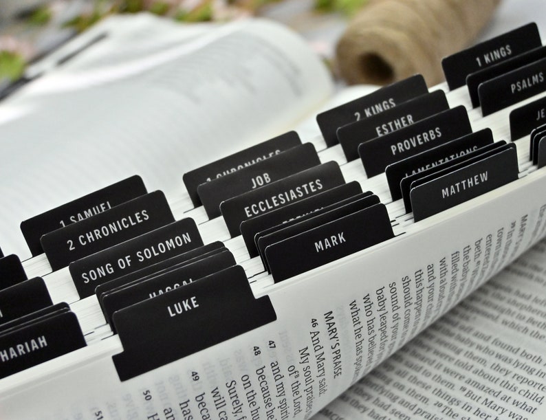 May include: Close-up of a black and white Bible with black tabs marking each book of the Bible. The tabs are labeled with the names of the books, including "Song of Solomon", "Mark", "Matthew", "Esther", "Proverbs", and "1 Kings".