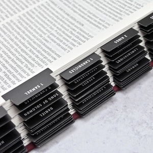 May include: A close-up of a black and white Bible with black tabs marking each book of the Bible. The tabs are labeled with the names of the books, such as "1 Samuel", "2 Samuel", "1 Kings", "2 Kings", "1 Chronicles", "2 Chronicles", "Daniel", "Song of Solomon", "Ecclesiastes", "Ezekiel", "Jeremiah", "Lamentations", "Romans", "1 Corinthians", "Hosea", and "Isaiah".