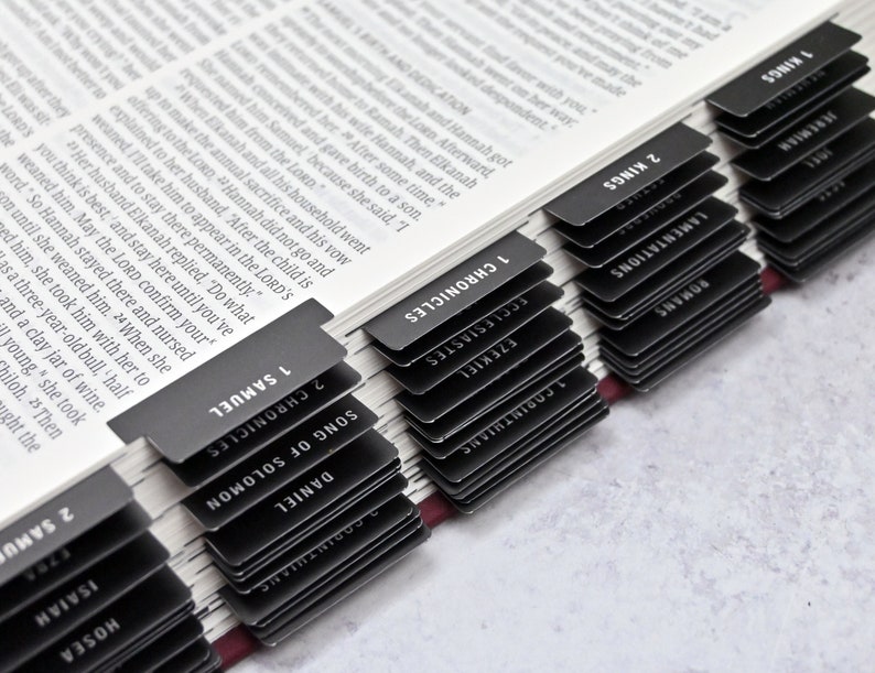 May include: A close-up of a black and white Bible with black tabs marking each book of the Bible. The tabs are labeled with the names of the books, such as "1 Samuel", "2 Samuel", "1 Kings", "2 Kings", "1 Chronicles", "2 Chronicles", "Daniel", "Song of Solomon", "Ecclesiastes", "Ezekiel", "Jeremiah", "Lamentations", "Romans", "1 Corinthians", "Hosea", and "Isaiah".