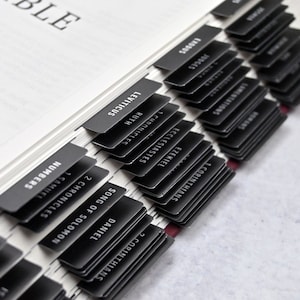 May include: Close-up of a black and white Bible with black tabs for each book of the Bible. The tabs are labeled with the names of the books, including Numbers, 1 Chronicles, 2 Chronicles, Song of Solomon, Daniel, 1 Corinthians, 2 Corinthians, Lamentations, Ezekiel, Ecclesiastes, Ruth, 1 Samuel, Leviticus, Exodus, Judges, and more.