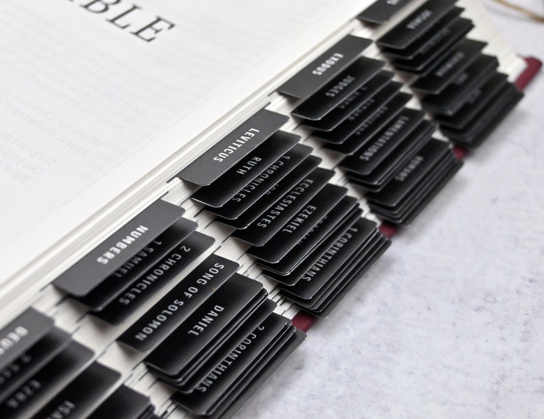 May include: Close-up of a black and white Bible with black tabs for each book of the Bible. The tabs are labeled with the names of the books, including Numbers, 1 Chronicles, 2 Chronicles, Song of Solomon, Daniel, 1 Corinthians, 2 Corinthians, Lamentations, Ezekiel, Ecclesiastes, Ruth, 1 Samuel, Leviticus, Exodus, Judges, and more.
