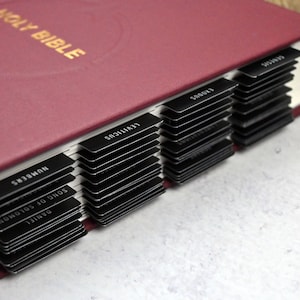 May include: A burgundy leather-bound Holy Bible with black tabs for each book of the Bible. The tabs are labeled with the book names, including "Daniel", "Song of Solomon", "Numbers", "Leviticus", "Exodus", and "Genesis".