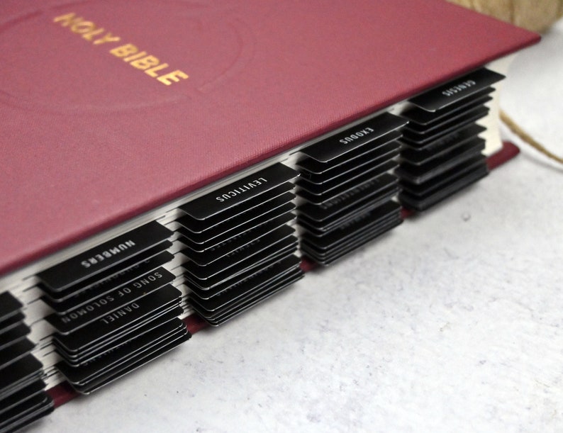May include: A burgundy leather-bound Holy Bible with black tabs for each book of the Bible. The tabs are labeled with the book names, including "Daniel", "Song of Solomon", "Numbers", "Leviticus", "Exodus", and "Genesis".