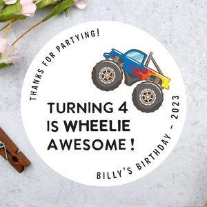May include: A white circular sticker with a cartoon monster truck design. The truck is blue, red, and yellow. Text reads "Turning 4 is Wheelie Awesome!" and "Billy's Birthday - 2023".