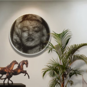 Puede incluir: Una pieza de arte de pared circular con un diseño de rostro en hilo marrón. El arte está sobre un fondo blanco y está colgado en una pared blanca. Una planta en maceta y dos esculturas de caballos de bronce están en primer plano.