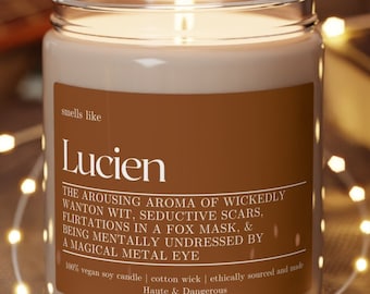 May include: A brown candle with a white label that reads "smells like Lucien" and a description of the scent. The candle is on a wooden coaster and surrounded by string lights.