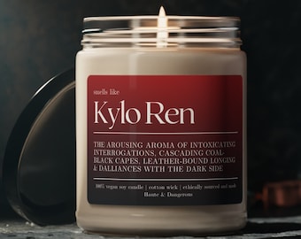 May include: A white candle in a glass jar with a black label that reads "smells like Kylo Ren" and describes the scent as "The arousing aroma of intoxicating interrogations, cascading coal black capes, leather-bound longing & dalliances with the dark side". The label also states that the candle is 100% vegan soy candle, cotton wick, ethically sourced and made.
