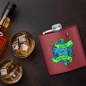 May include: A red metal flask with a blue and green illustration of a twenty-sided die, a staff, and the text "Selena Sorcerer 20". The flask is on a brown surface with two glasses of ice water.