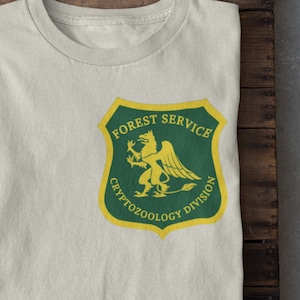 May include: A white t-shirt with a green and yellow patch that reads "Forest Service Cryptozoology Division". The patch features a yellow mythical creature with wings.