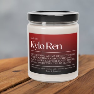 May include: A white candle jar with a black lid. The label is red and says "smells like Kylo Ren" and "The Arousing Aroma of Intoxicating Interrogations. Cascading Coal-Black Capes. Leather-Bound Longing & Dalliances With The Dark Side". The label also says "100% vegan soy candle | cotton wick | ethically sourced and made Haute & Dangerous."